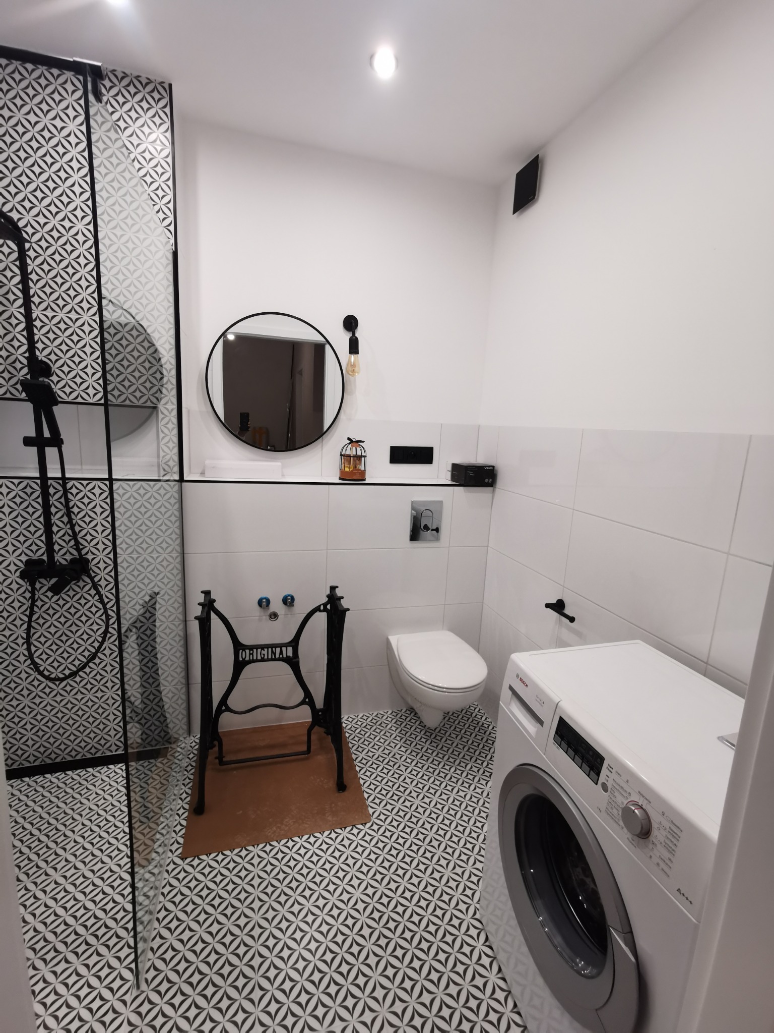 Nowoczesna łazienka z kabiną prysznicową typu walk-in, umywalką na stelażu maszyny do szycia Singer i pralką Bosch. Biało-czarna kolorystyka, wzorzyste płytki podłogowe.