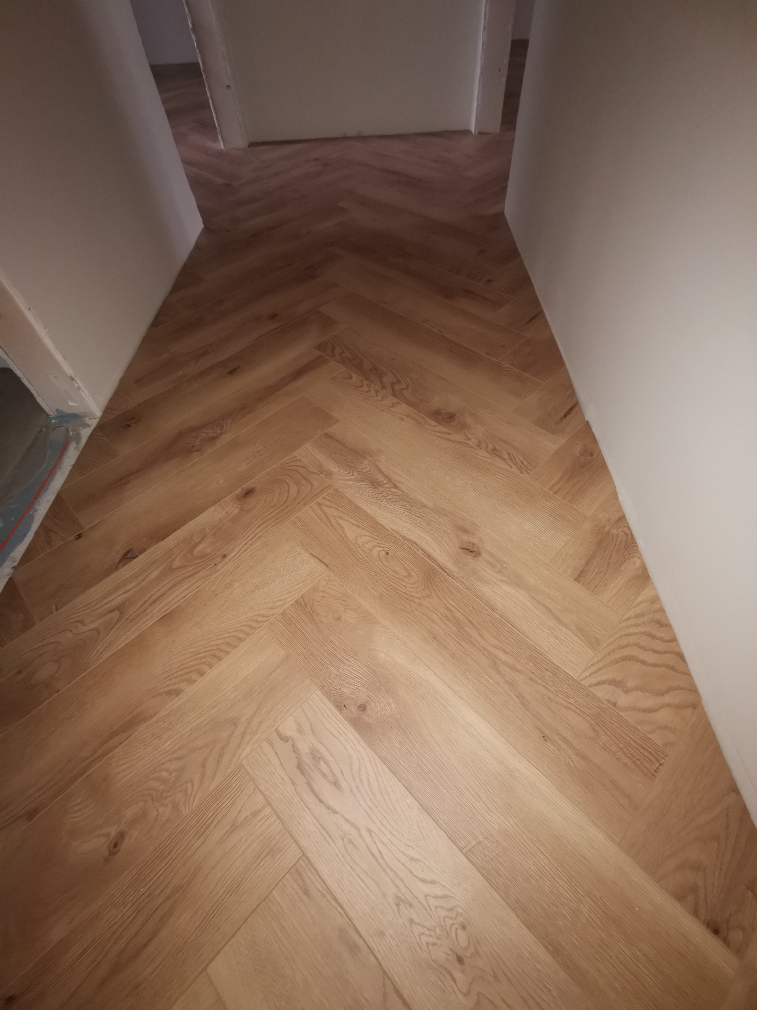 Ułożona podłoga z paneli w jodełkę, widok z góry, jasne drewno, białe ściany, perspektywa w głąb korytarza. Widoczne detale łączeń paneli i struktura drewna.