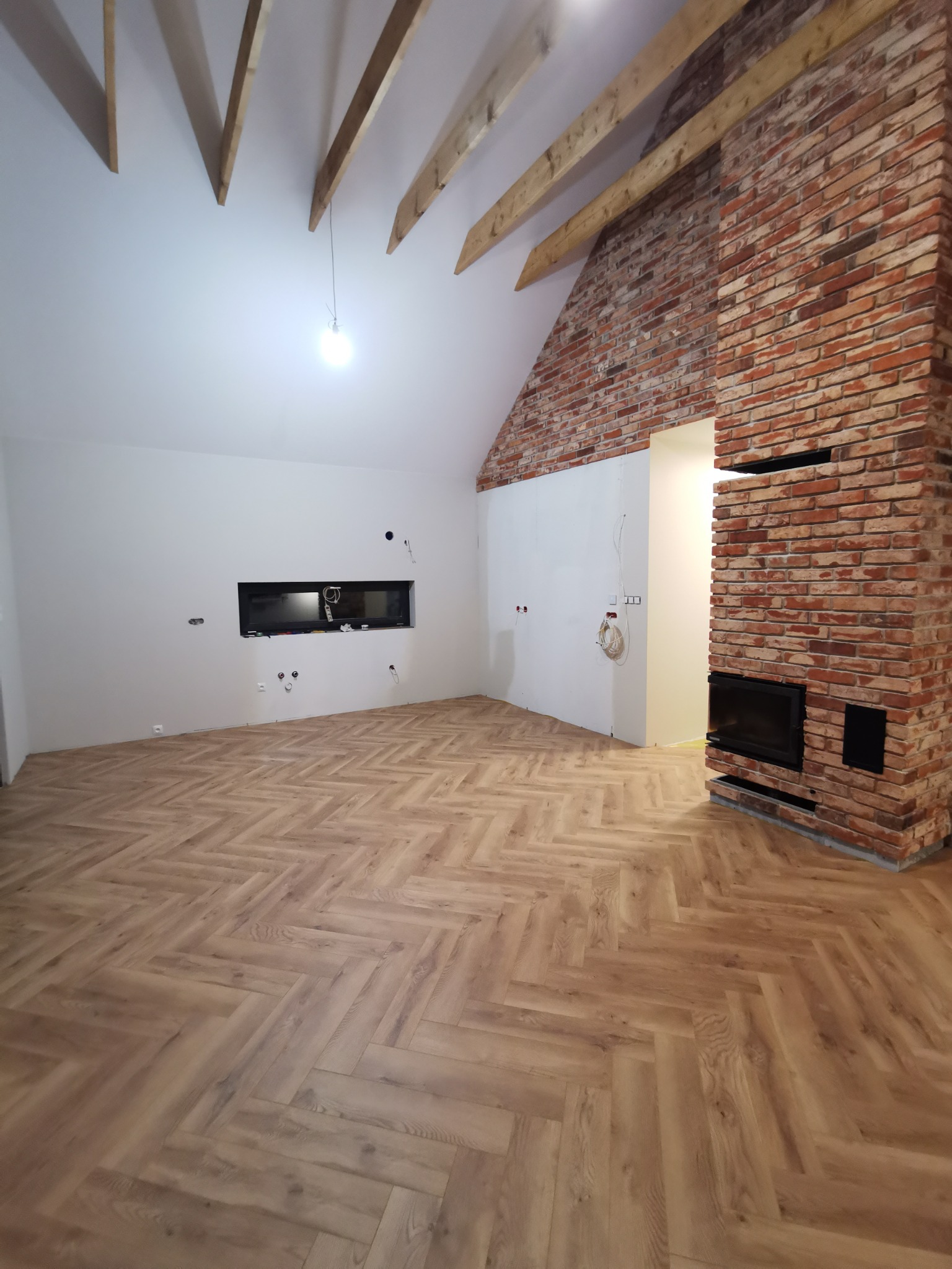 Salon z parkietem w jodełkę, ceglanym kominkiem i surowym drewnem na suficie. Widoczne przygotowania do montażu oświetlenia i gniazdek. Wnętrze w trakcie wykańczania.