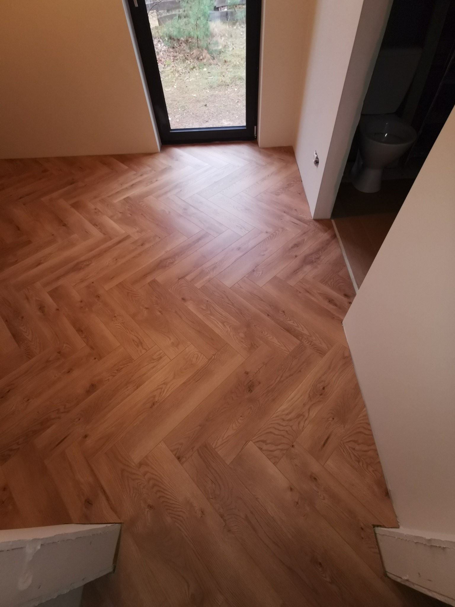 Ułożona podłoga z paneli w jodełkę w jasnym odcieniu drewna, widok z góry na fragment łazienki z toaletą w tle, okno z widokiem na zewnątrz.