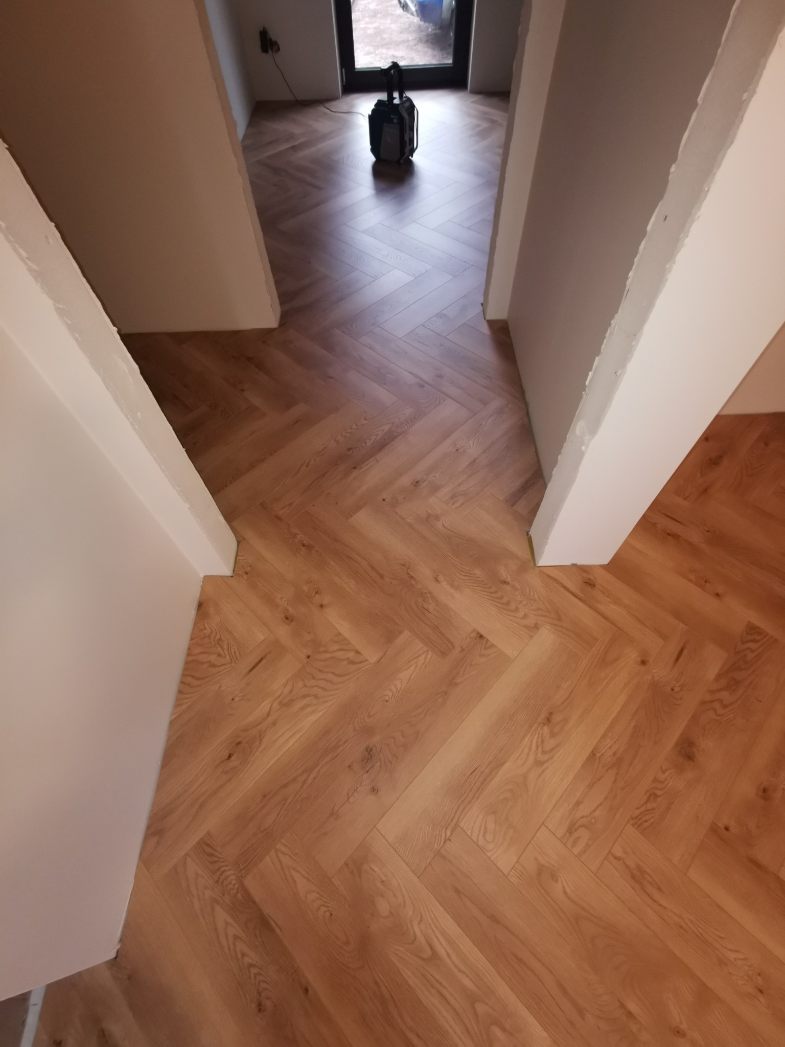 Ułożona parkietowa podłoga w jodełkę, widok z góry, z fragmentem białej ściany i przejściem do kolejnego pomieszczenia. W tle widoczny odkurzacz.