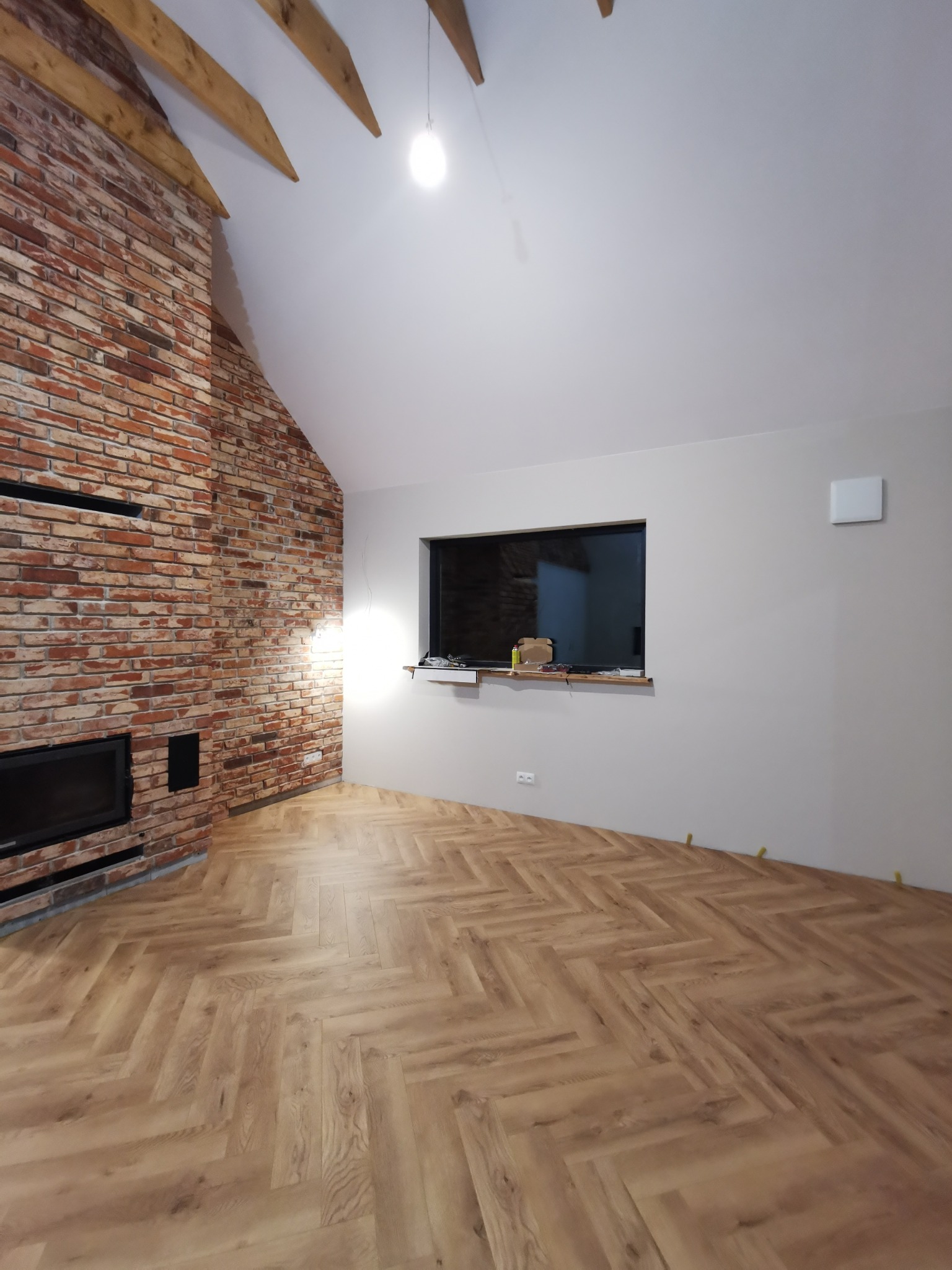Salon z ceglaną ścianą kominkową i parkietem ułożonym w jodełkę. Widoczne drewniane belki stropowe i okno z widokiem na ceglaną ścianę. Wnętrze w trakcie wykańczania.