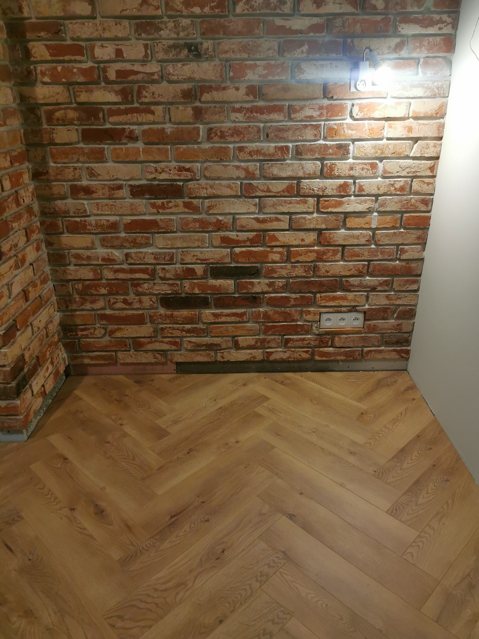 Ukończona ściana z cegły i parkiet ułożony w jodełkę. Widoczne gniazdka elektryczne i oświetlenie punktowe. Detal wykończenia wnętrza.