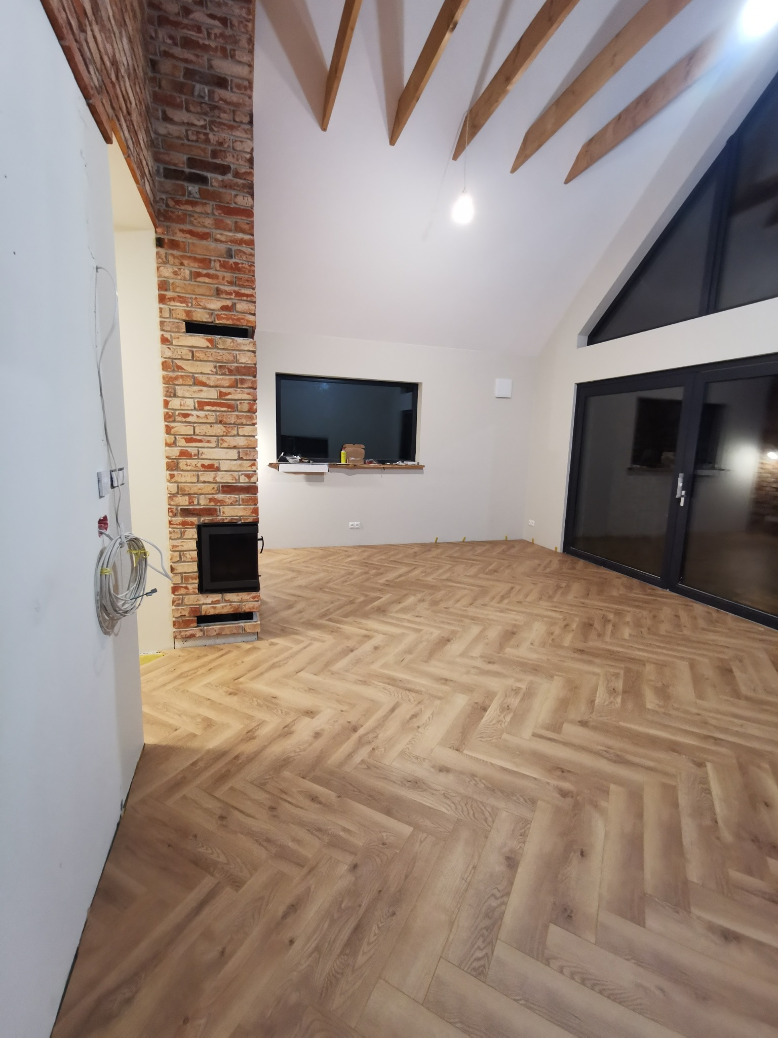 Salon z nową podłogą w jodełkę, ceglanym kominem i surowym drewnem na suficie. Widoczne okno i drzwi balkonowe. Kabel wystaje ze ściany.