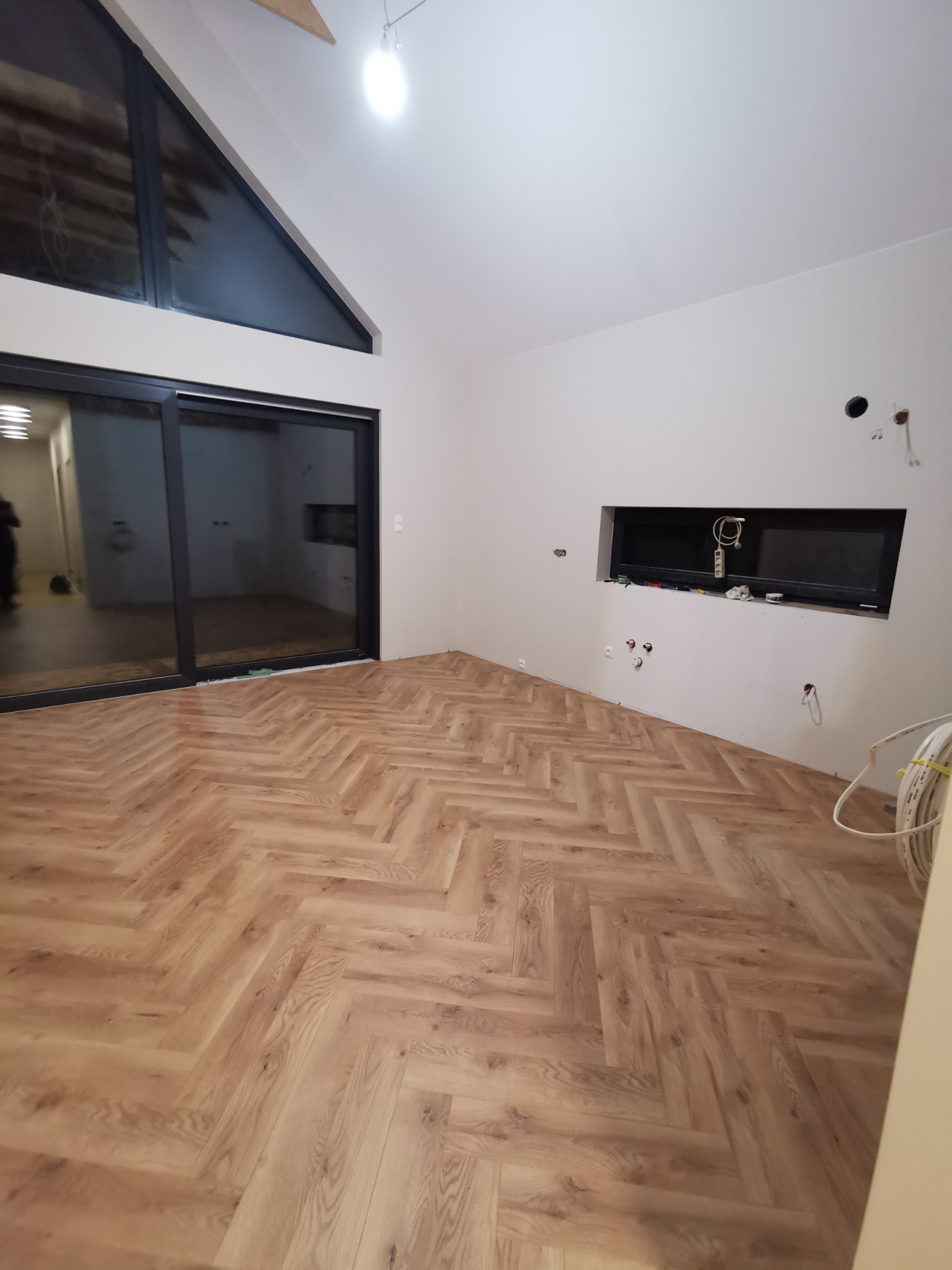 Ułożona podłoga z paneli w jodełkę w jasnym, nowoczesnym wnętrzu z dużym oknem i przygotowanym miejscem na telewizor. Widoczne kable i instalacje.