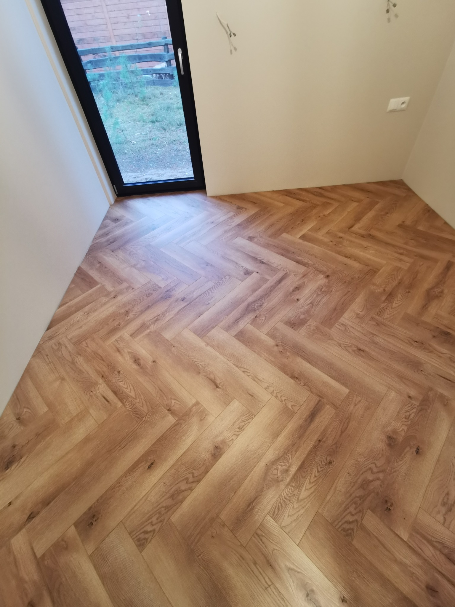 Ukończona podłoga z paneli ułożonych w jodełkę, widok z góry. Jasne ściany, czarna rama okienna. Widoczne gniazdko elektryczne i fragment instalacji oświetleniowej.