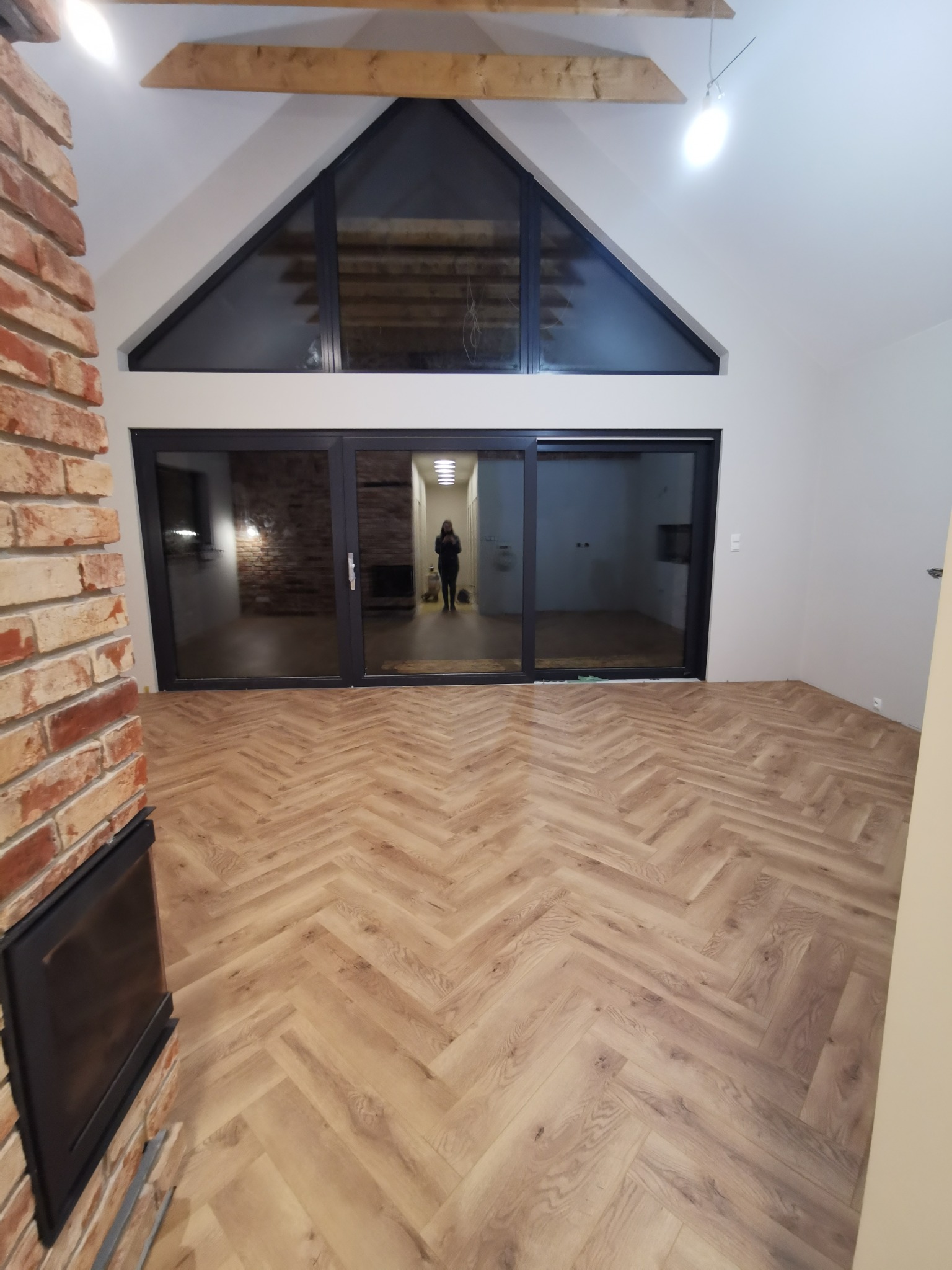 Jasne panele ułożone w jodełkę francuską w przestronnym salonie z ceglanym kominkiem i dużym oknem. Widoczne drewniane belki stropowe i przesuwne drzwi tarasowe.