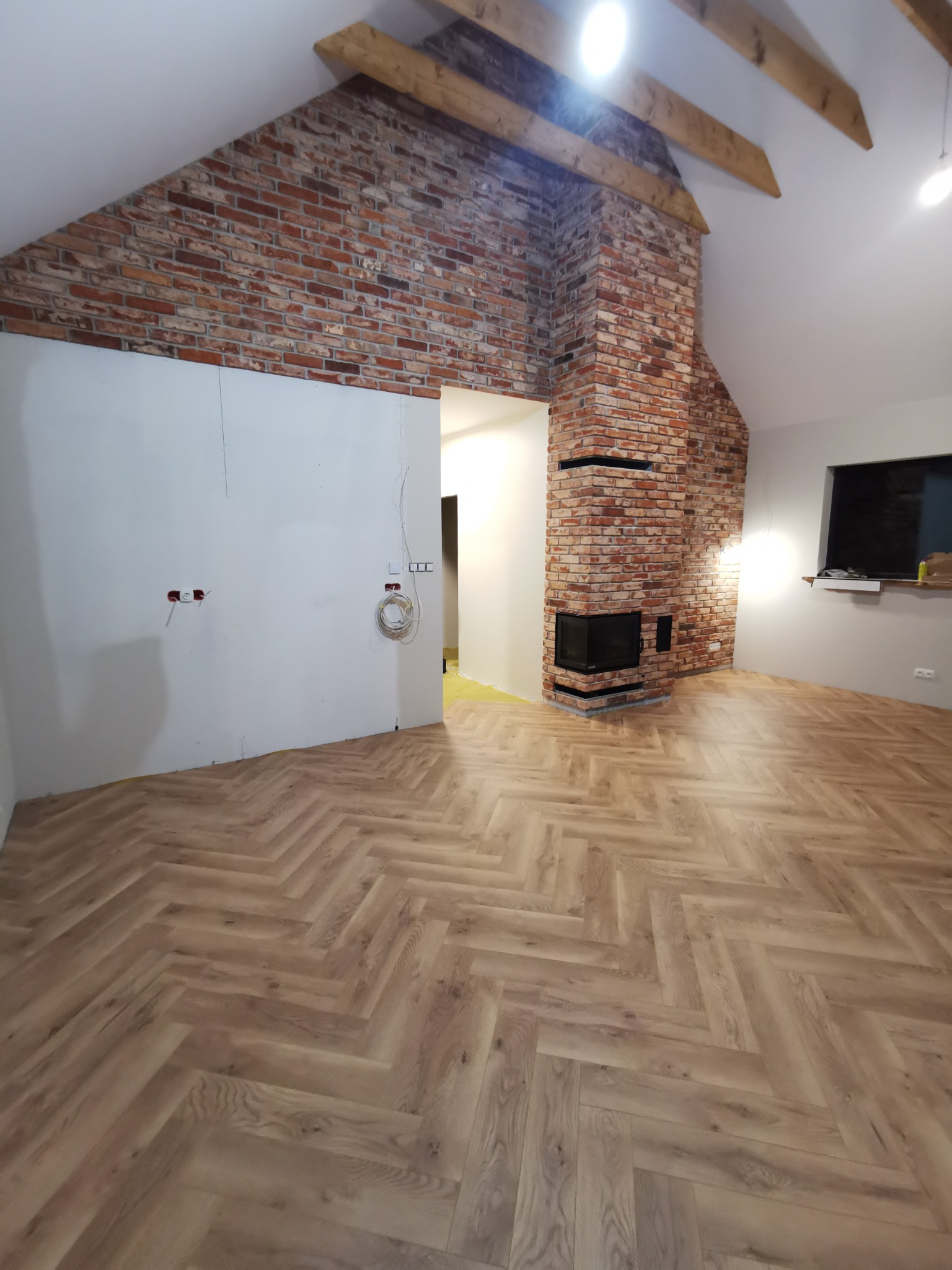 Ukończone układanie parkietu w jodełkę w pokoju z ceglanym kominkiem i surowymi belkami. Częściowo wykończone ściany, widoczne gniazdka elektryczne.