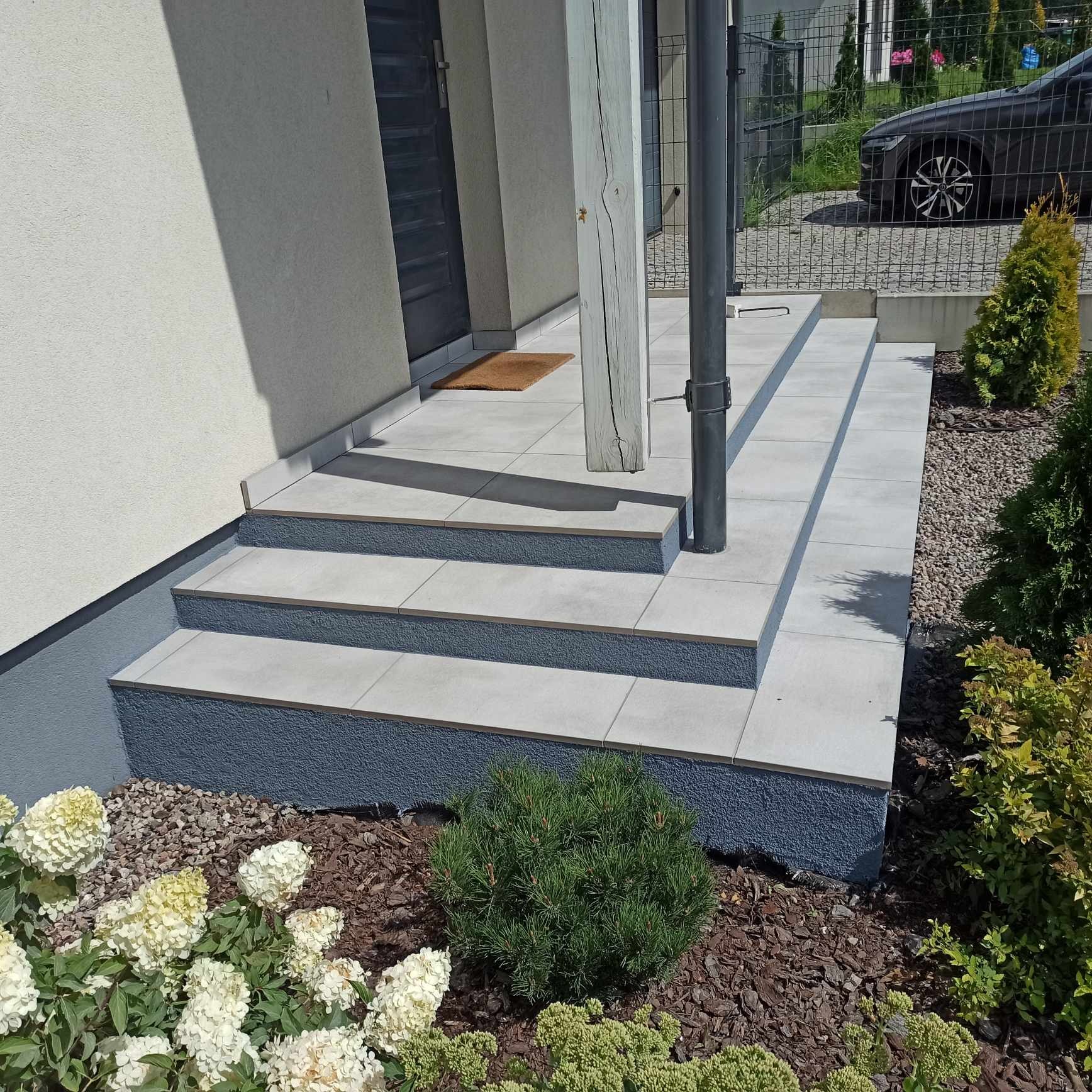Wykończone schody wejściowe do domu z szarych płytek, z ciemnoszarą podmurówką i elementami architektury ogrodowej. Widok na drzwi wejściowe i fragment podjazdu.