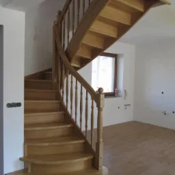 Kręte, jasne schody z drewna z toczonymi balustradami w jasnym wnętrzu. Widoczne okno z żaluzjami i jasna podłoga. Detal wykończenia.