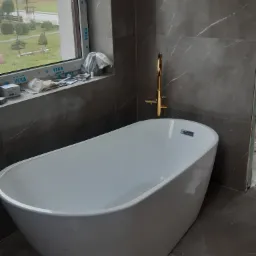 Nowoczesna, wolnostojąca wanna z złotą armaturą na tle ściany wyłożonej szarymi płytkami imitującymi marmur, widok na okno z widokiem na zewnątrz, elementy wykończeniowe wciąż widoczne.