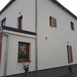 Wykończona elewacja domu jednorodzinnego z tynkiem w odcieniach bieli i szarości, z drewnopodobną okładziną fragmentu ściany i okiennicami, widoczne rynny i oświetlenie zewnętrzne.