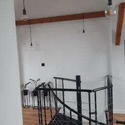 Wnętrze z drewnianymi belkami stropowymi, wiszącymi żarówkami Edison, spiralnymi schodami z czarnego metalu i roślinami doniczkowymi w minimalistycznych, czarnych stojakach.