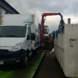 Biały samochód dostawczy Iveco z otwartą skrzynią ładunkową stoi obok kontenera na gruz, w tle widoczna koparka z chwytakiem hydraulicznym załadowująca odpady do kontenera, budynek z oknami w tle.