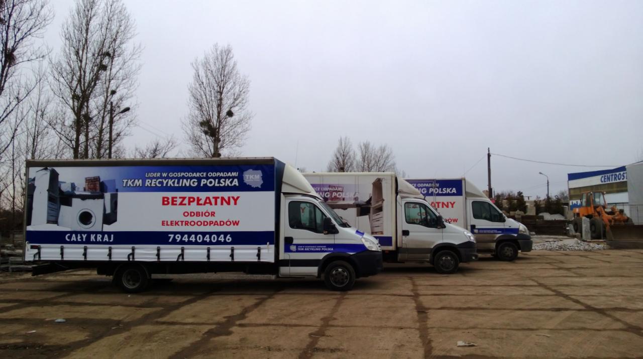 Trzy białe samochody dostawcze z niebieskimi napisami 'TKM Recykling Polska' i informacją o bezpłatnym odbiorze elektroodpadów, stojące na placu, w tle budynek z napisem 'Centros' i pomarańczowa...