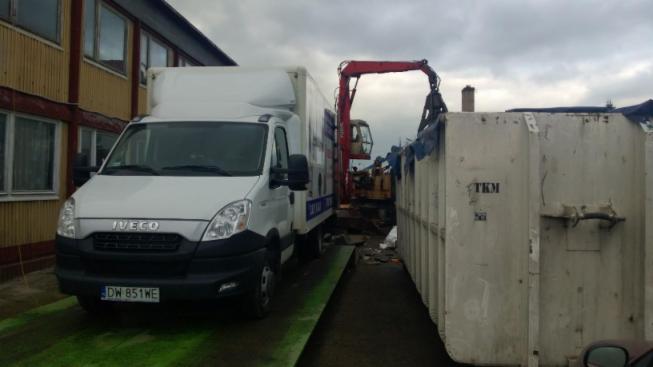 Biały samochód dostawczy Iveco z otwartą skrzynią ładunkową stoi obok kontenera na gruz, w tle widoczna koparka z chwytakiem hydraulicznym załadowująca odpady do kontenera, budynek z oknami w tle.