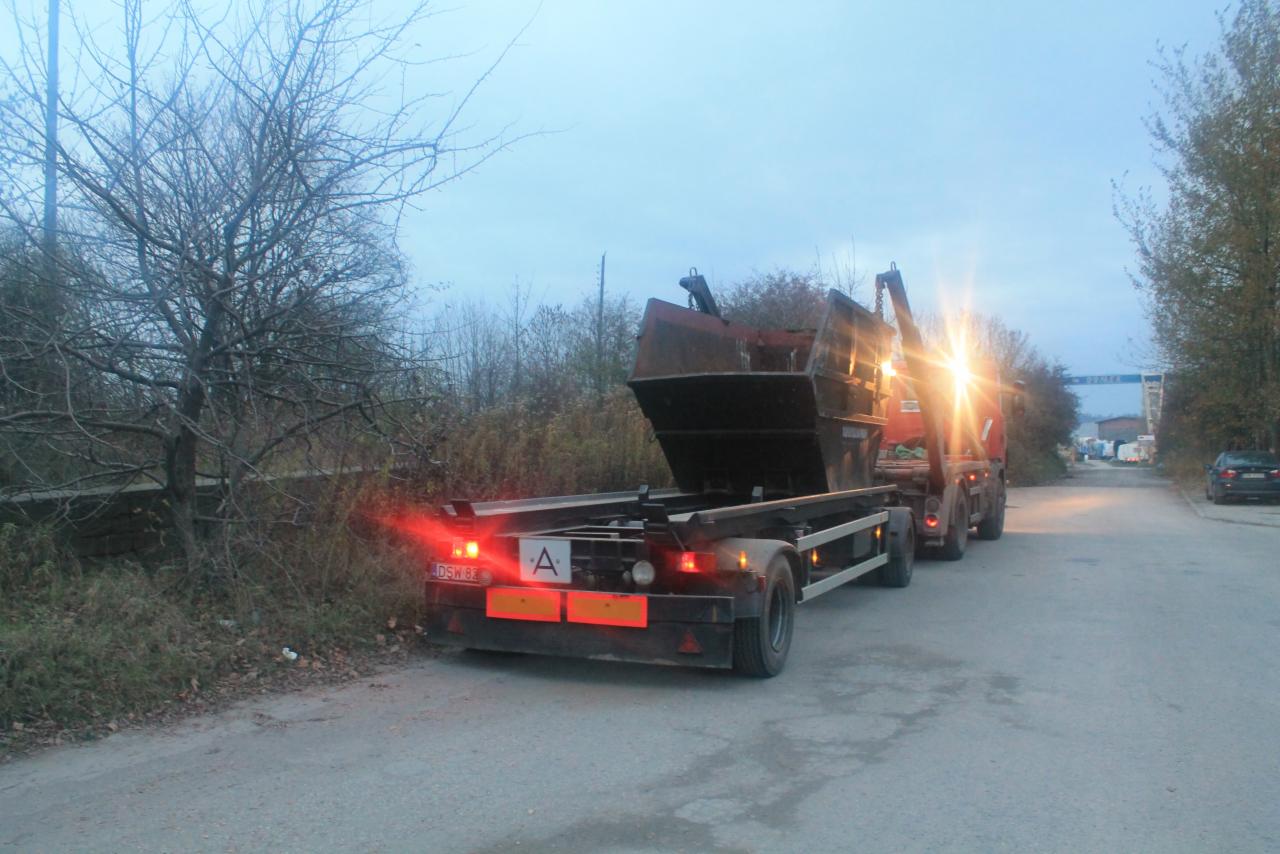 Ciężarówka hakowa z otwartym kontenerem na poboczu drogi w pochmurny dzień, widoczne światła stopu i odblaski, drzewa bez liści w tle.