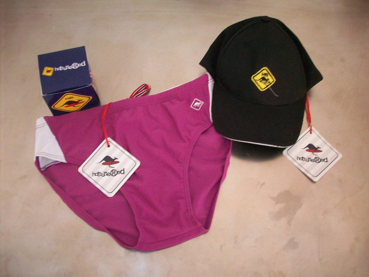 Fioletowe damskie figi z białymi wstawkami, czarna czapka z daszkiem z żółtym logo i pudełko z logo marki Hot Buttered, prezentowane na beżowym tle.