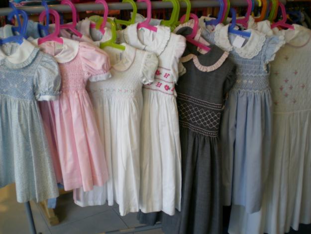 Rząd używanych sukienek dla dziewczynek w pastelowych kolorach, wiszących na różnokolorowych plastikowych wieszakach, z detalami smockingowymi i kołnierzykami.