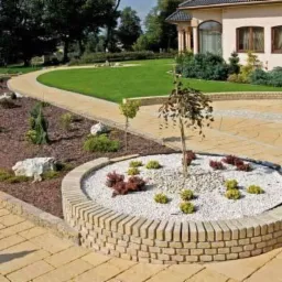 Żółta kostka brukowa tworząca ścieżkę i podjazd, obrzeża rabat z brązową korą, biały żwir i rośliny w okrągłej rabacie, trawnik i dom w tle.