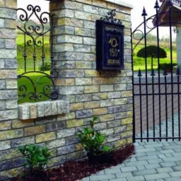 Murowane ogrodzenie z kamienia naturalnego z metalową bramą i skrzynką pocztową z numerem domu, otoczone kostką brukową i roślinami.