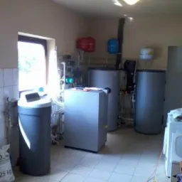 Wnętrze kotłowni z zainstalowanymi urządzeniami grzewczymi: zbiorniki, filtry i rury. Widoczne okno i płytki na podłodze. Niebieski i czerwony osprzęt na ścianie.