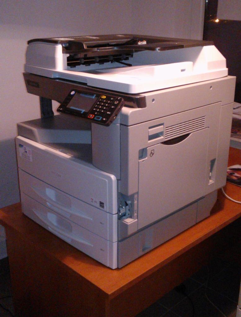 Nowa, szara kserokopiarka Ricoh Aficio MP 2001L formatu A3, umieszczona na biurku z drewna, widoczny panel sterowania i podajnik papieru.