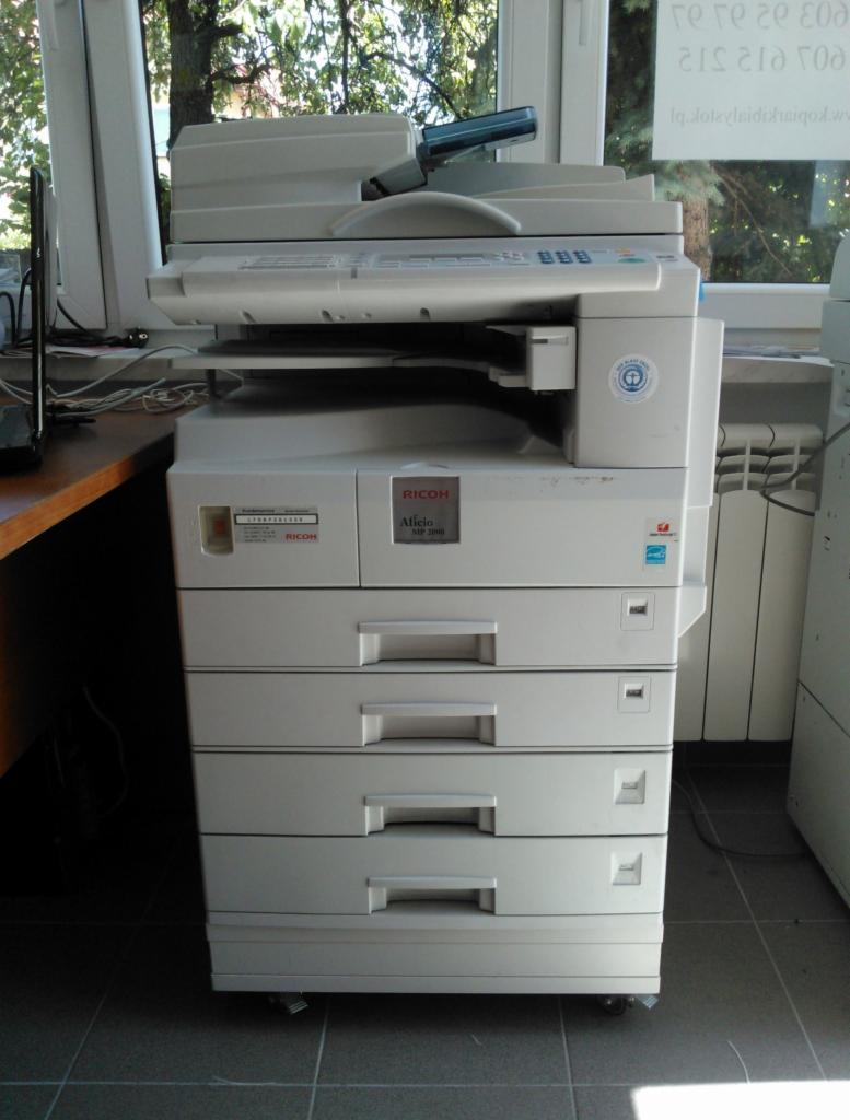 Kserokopiarka Ricoh Aficio MP 2000 z czterema szufladami na papier, widoczna w biurowym otoczeniu z oknem i zielenią w tle.