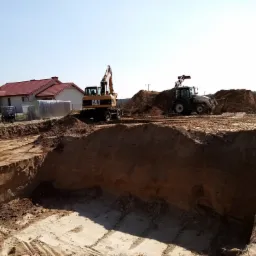 Wykop fundamentowy w piasku, z widocznymi śladami opon, koparka CAT i traktor z przodu, dom z czerwonym dachem w tle.