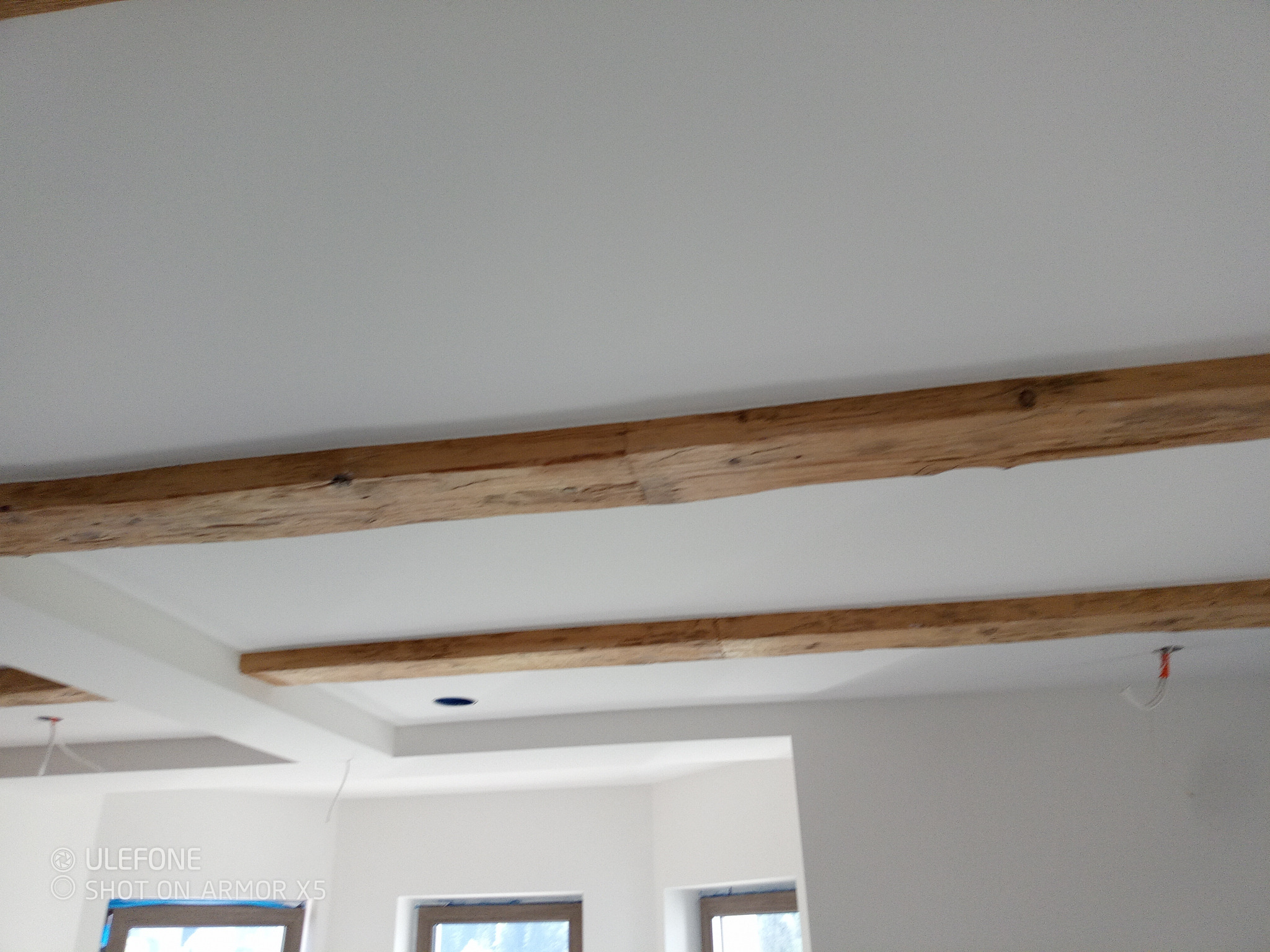 Jasne wnętrze z drewnianymi belkami stropowymi i podwieszanym sufitem. Widoczne okna i instalacja elektryczna. Minimalistyczny design.