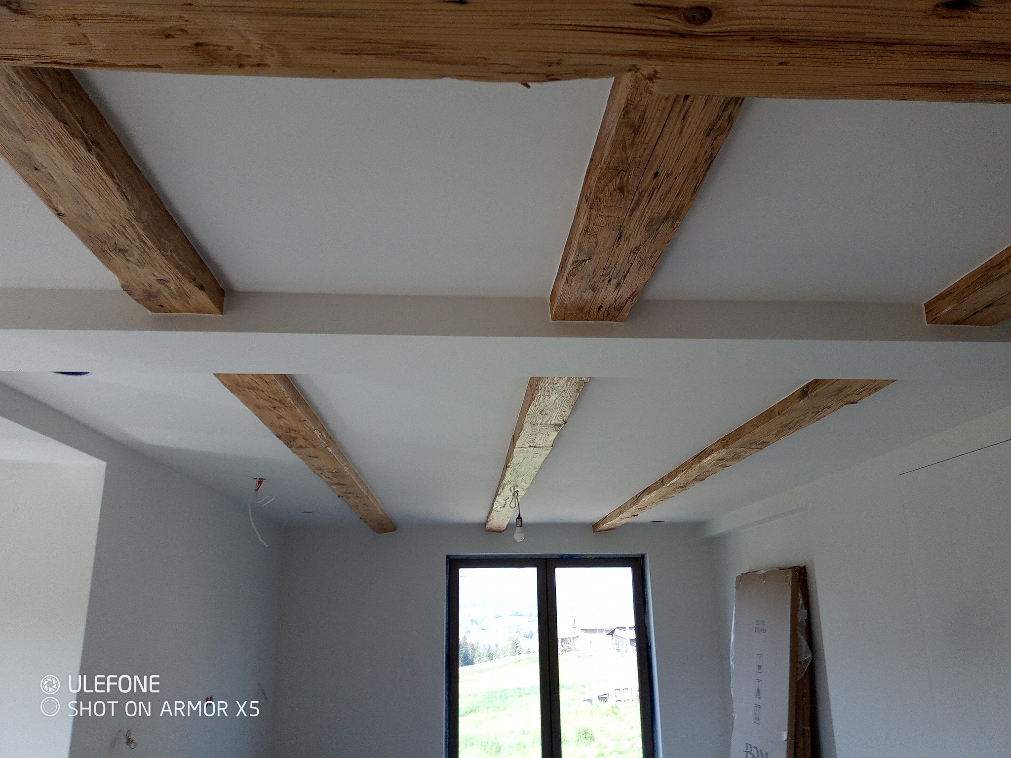 Białe wnętrze z drewnianymi belkami stropowymi, widok na okno z zielonym krajobrazem. Belki o surowym wykończeniu dodają rustykalnego charakteru.