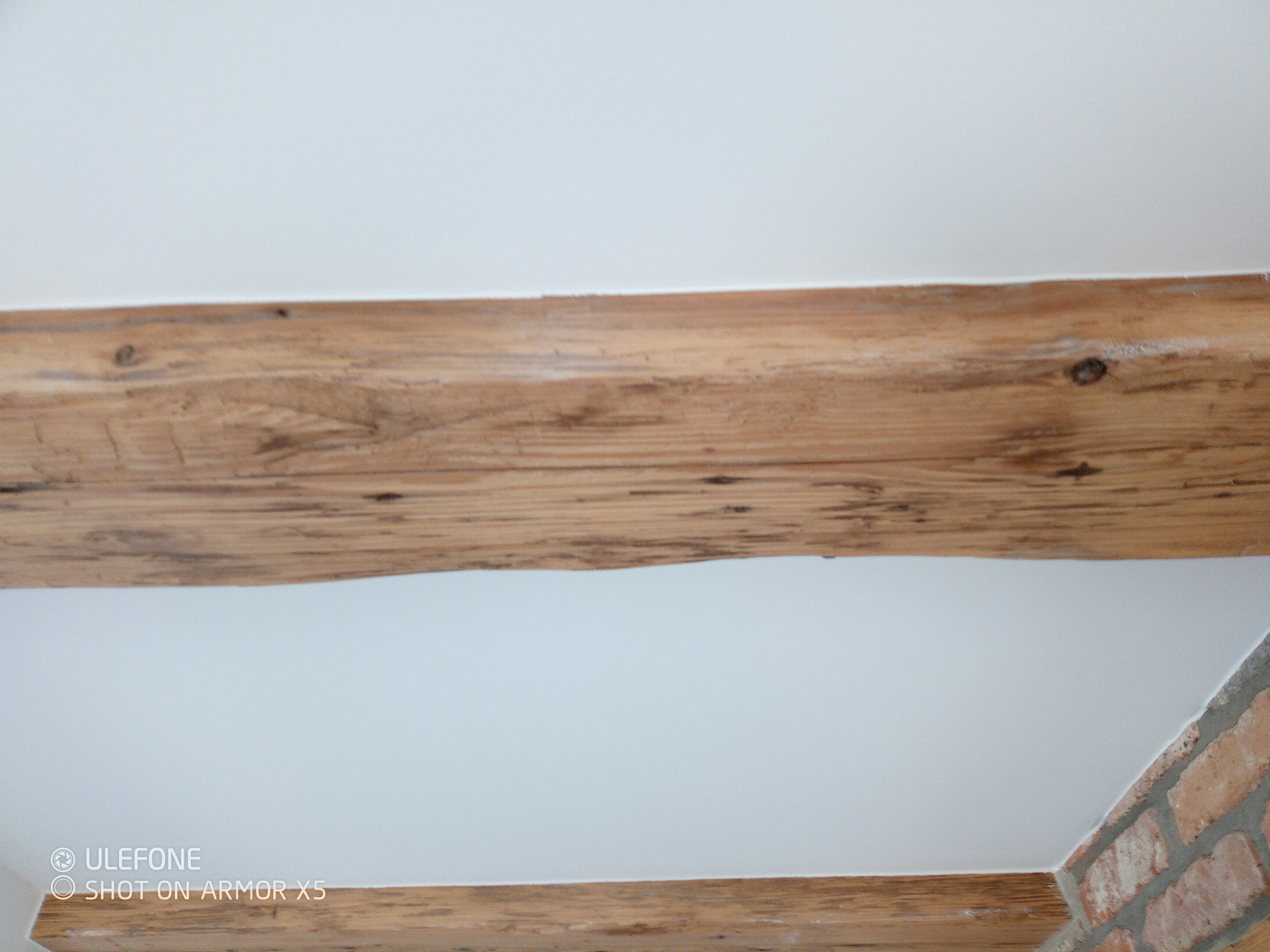 Drewniane belki stropowe z widoczną strukturą drewna na tle białego sufitu i fragmentu ściany z cegły. Ujęcie z dołu, podkreślające rustykalny charakter wnętrza.