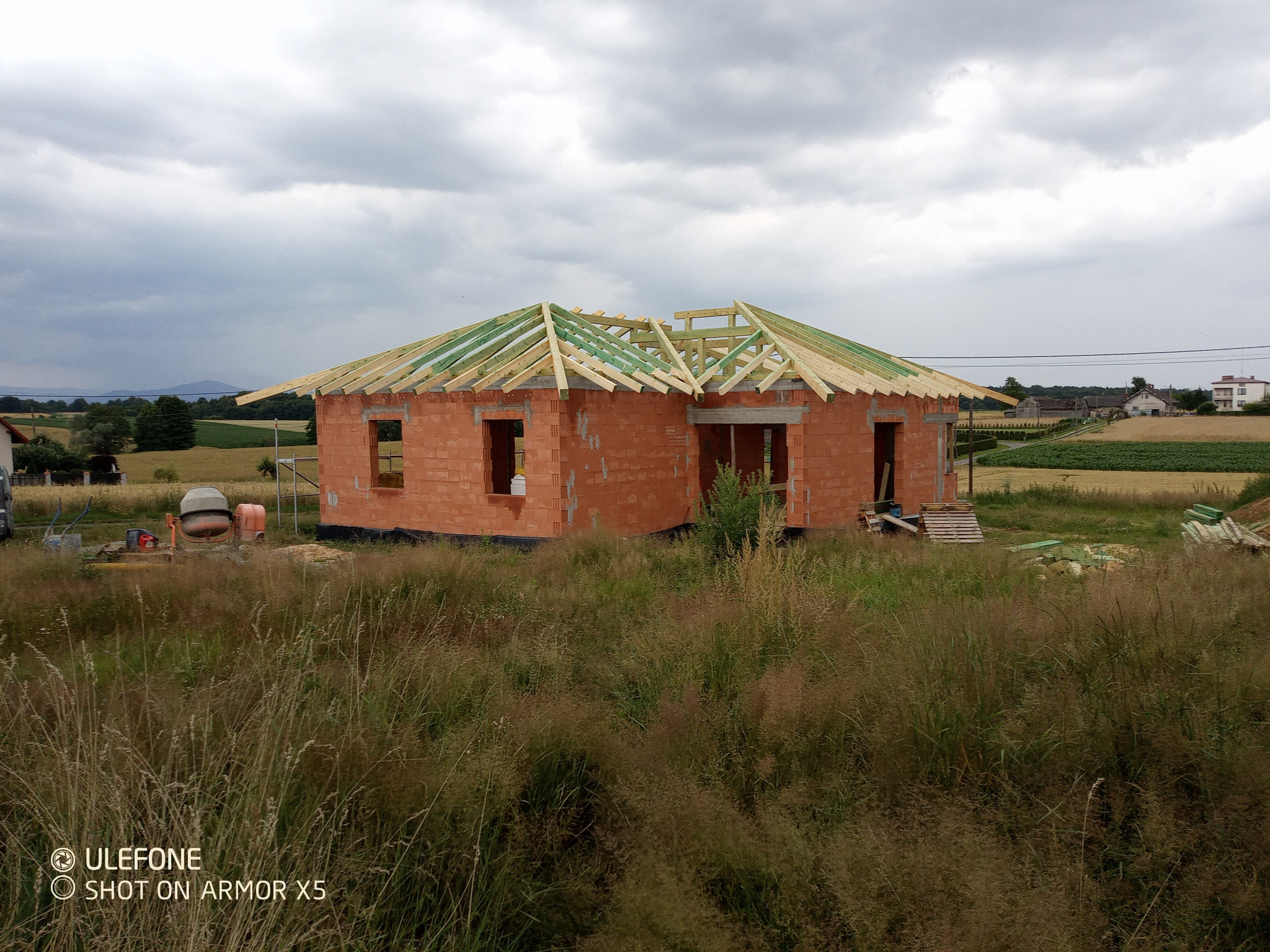 Niedokończony dom z czerwonej cegły z drewnianą konstrukcją dachu na tle zielonych pól i pochmurnego nieba. Widoczny mikser do betonu.