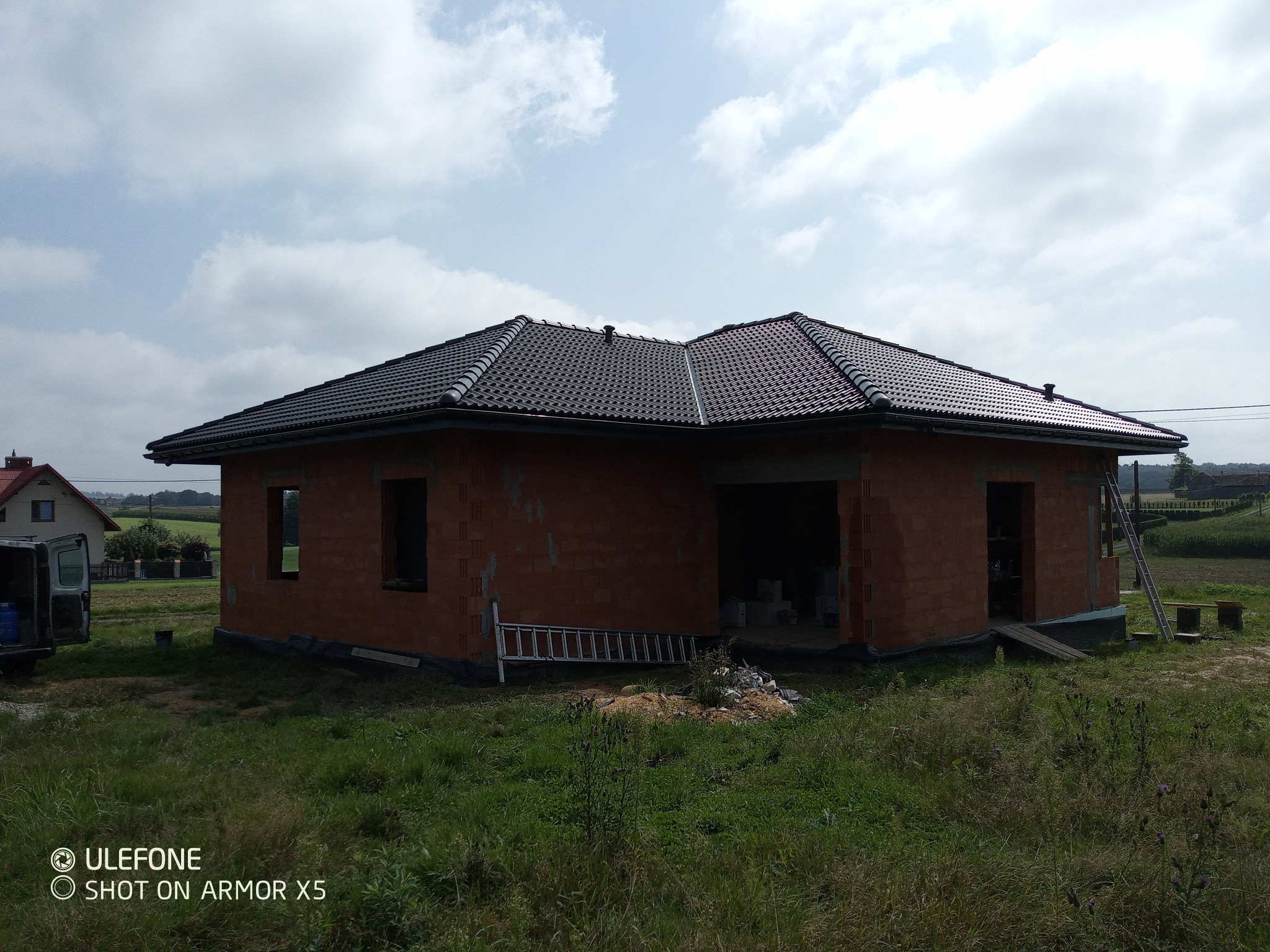 Niedokończony dom z czerwonej cegły z czarnym dachem na tle zielonego pola i błękitnego nieba. Widoczne rusztowania i drabina. Dom w stanie surowym zamkniętym.