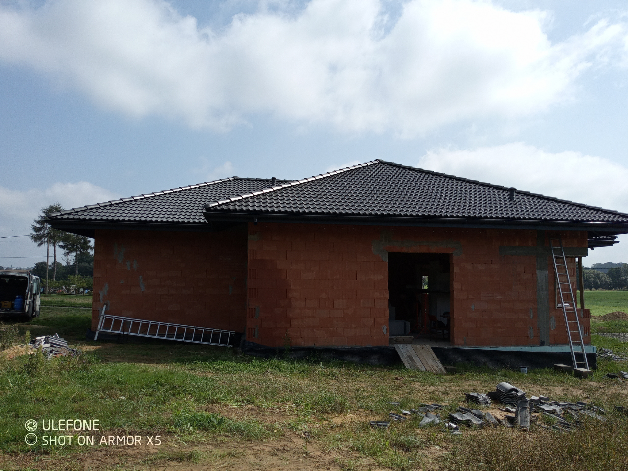 Surowy, jednopiętrowy dom z czerwonej cegły z czarnym dachem z dachówki, otoczony zielenią. Dwie drabiny opierają się o ściany. Widoczne fragmenty dachówek na ziemi.