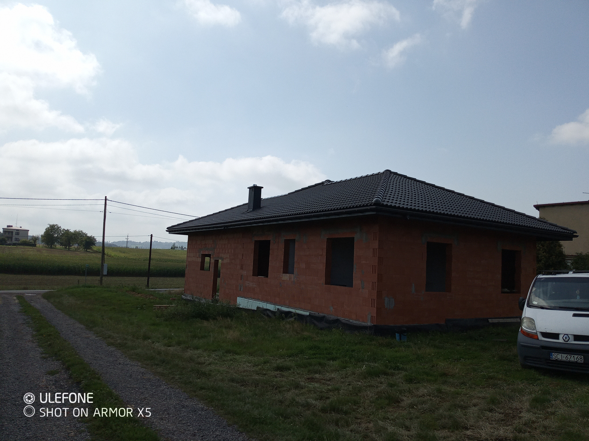 Surowy, jednopiętrowy dom z czerwonej cegły z czarnym dachem, w trakcie budowy, na tle zielonego pola i błękitnego nieba. Widoczny samochód dostawczy.