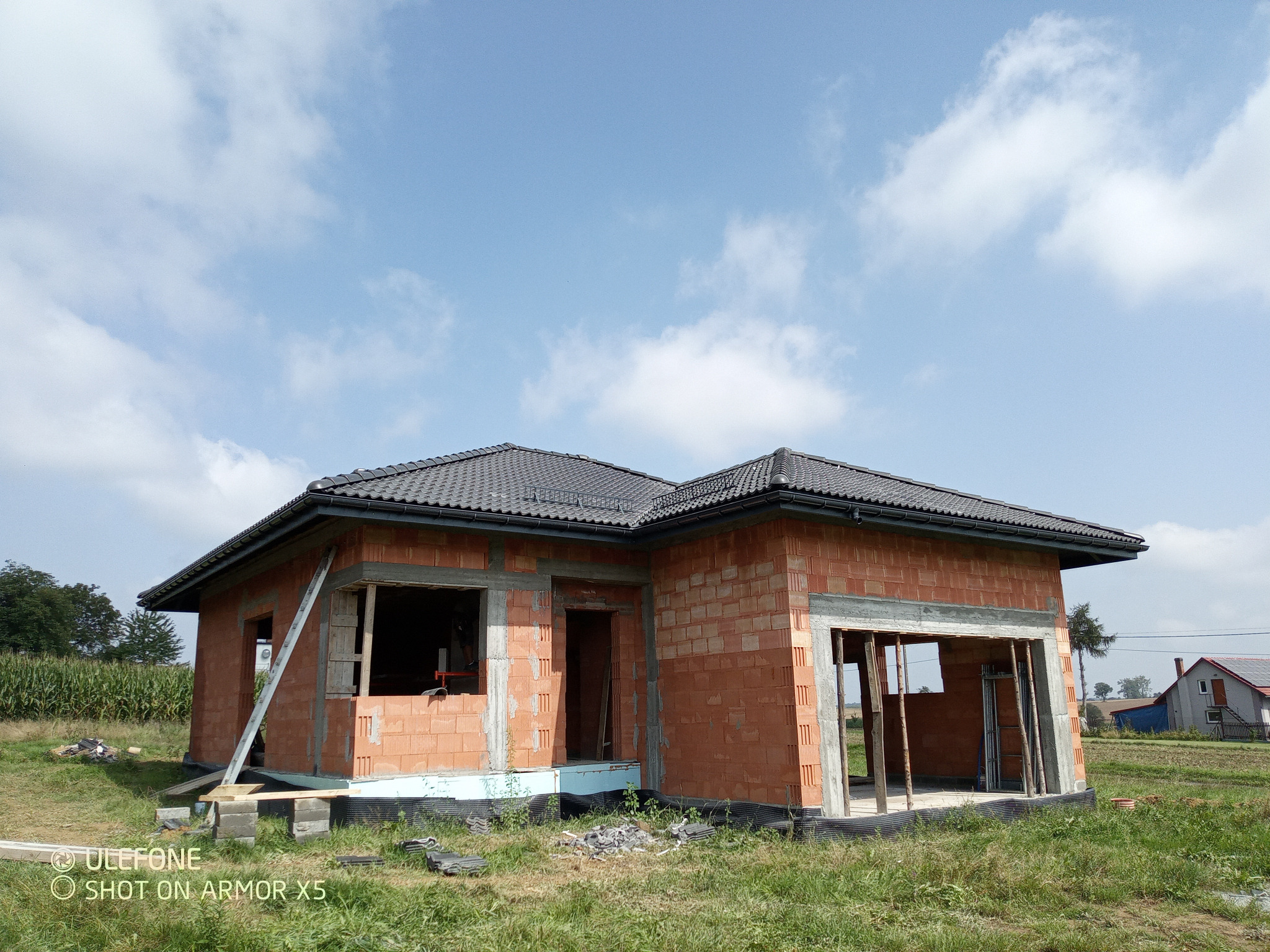 Niedokończony dom z czerwonej cegły z czarnym dachem na tle błękitnego nieba z chmurami. Widoczne otwory okienne i drzwiowe, teren trawiasty wokół.