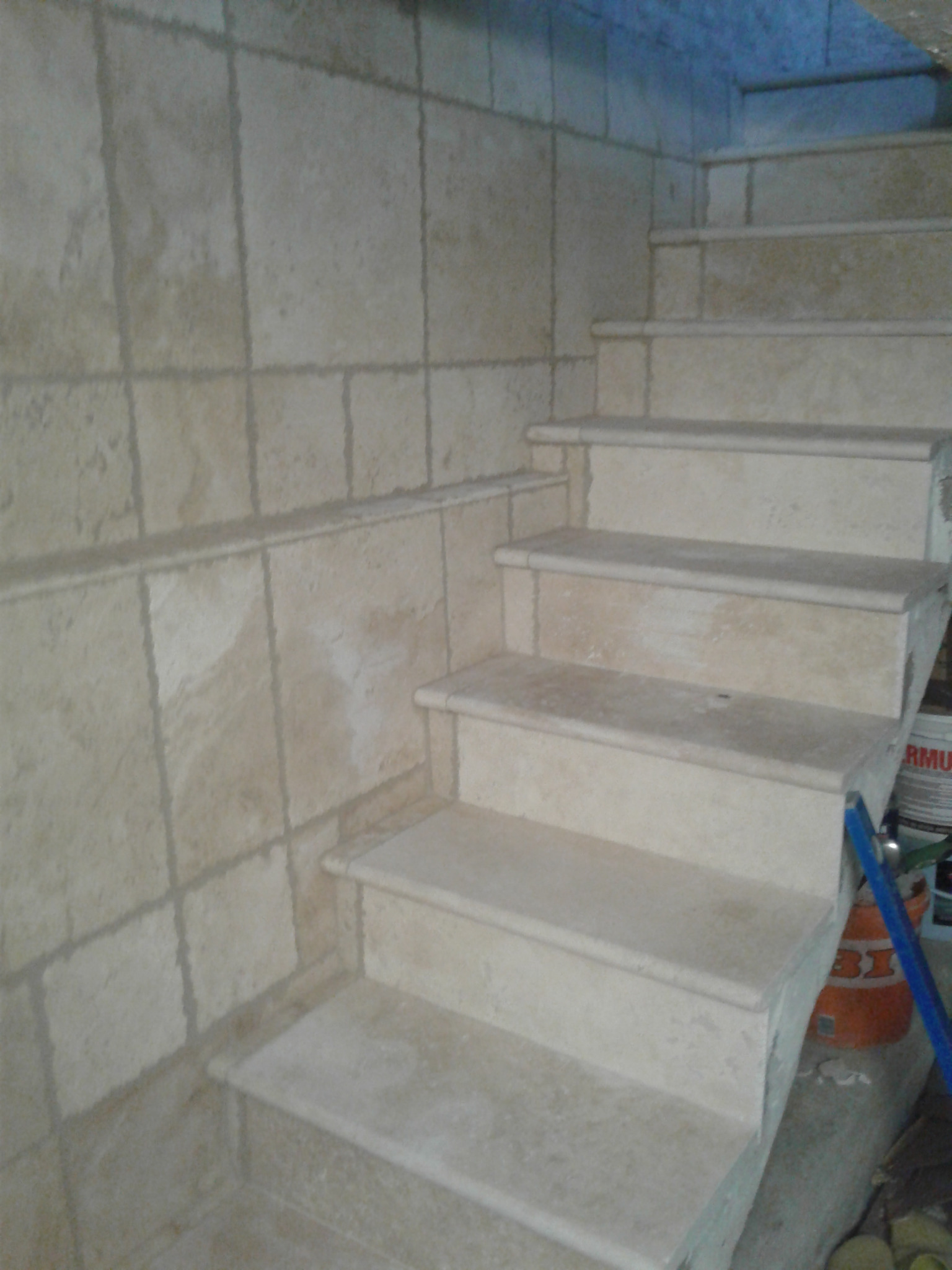 Wykończone schody wewnętrzne z beżowych płytek, z widocznymi fugami na ścianie. U dołu widoczne narzędzia i materiały budowlane. Ujęcie z dołu, perspektywa z poziomu podłogi.