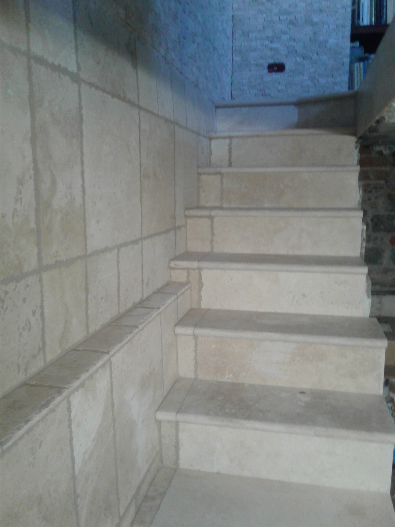 Wykończone schody z jasnego kamienia, z widocznymi fugami i okładziną ścienną z tego samego materiału. Ujęcie z dołu, podkreślające detale wykończenia.