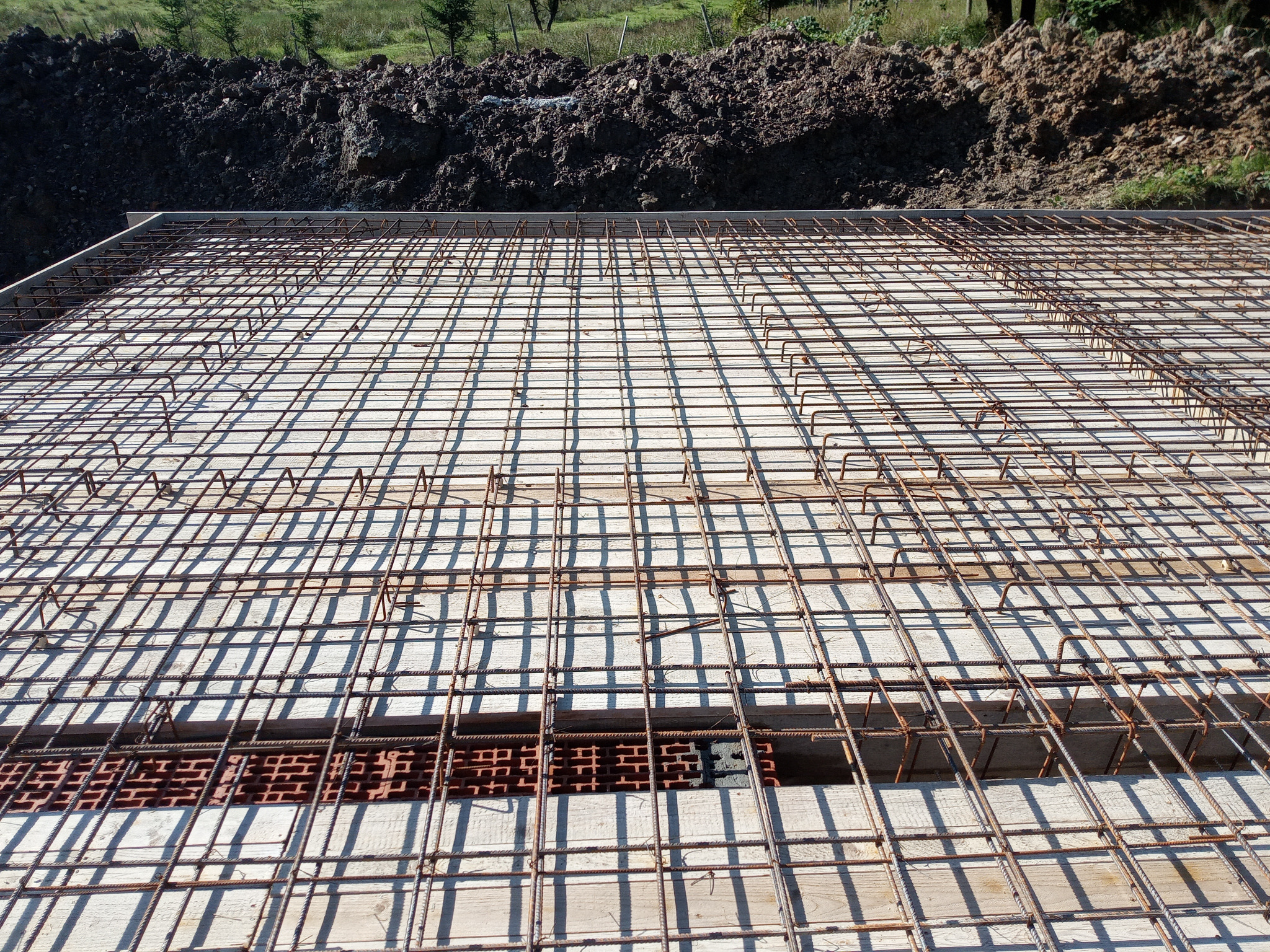 Zbrojenie płyty fundamentowej z widocznymi cegłami i deskowaniem, przygotowane do zalania betonem. W tle ziemia i zieleń.