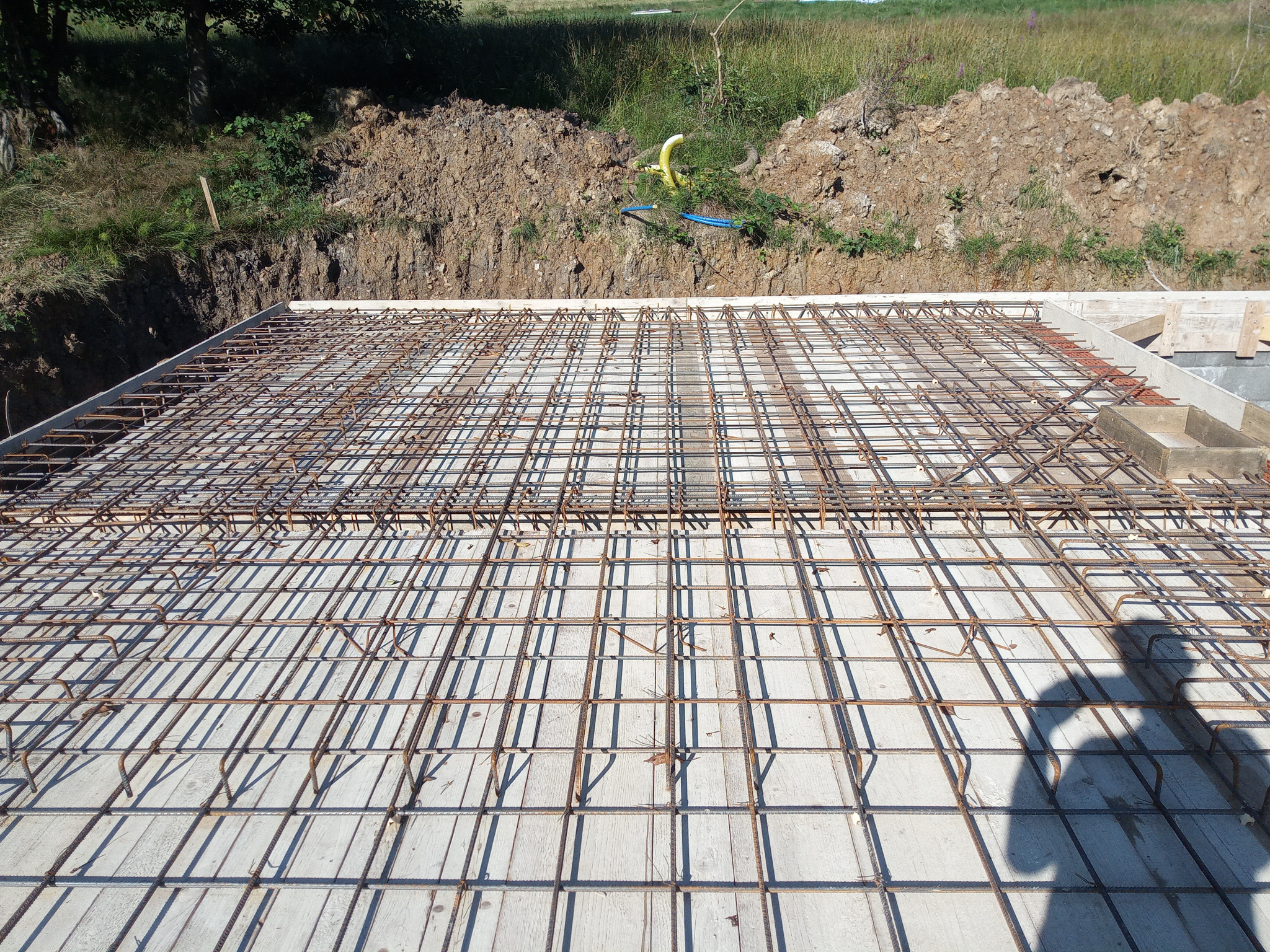 Zbrojenie płyty fundamentowej domu jednorodzinnego, widok z góry, zarys cienia osoby w prawym dolnym rogu. W tle ziemia i roślinność.