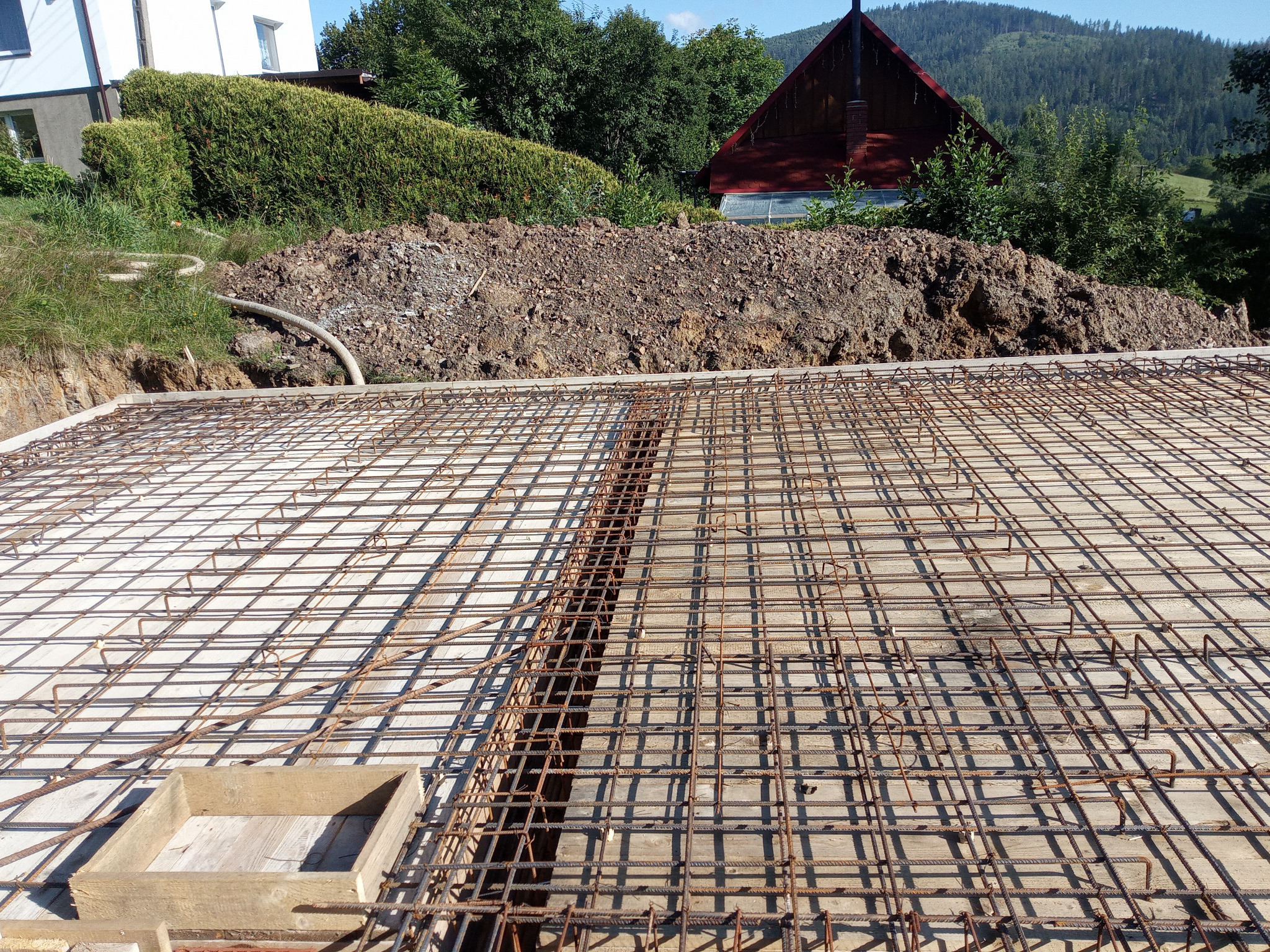 Zbrojenie fundamentów domu jednorodzinnego, widok z góry na siatkę z prętów stalowych, drewniany szalunek i ziemię w tle. Etap budowy w słoneczny dzień.