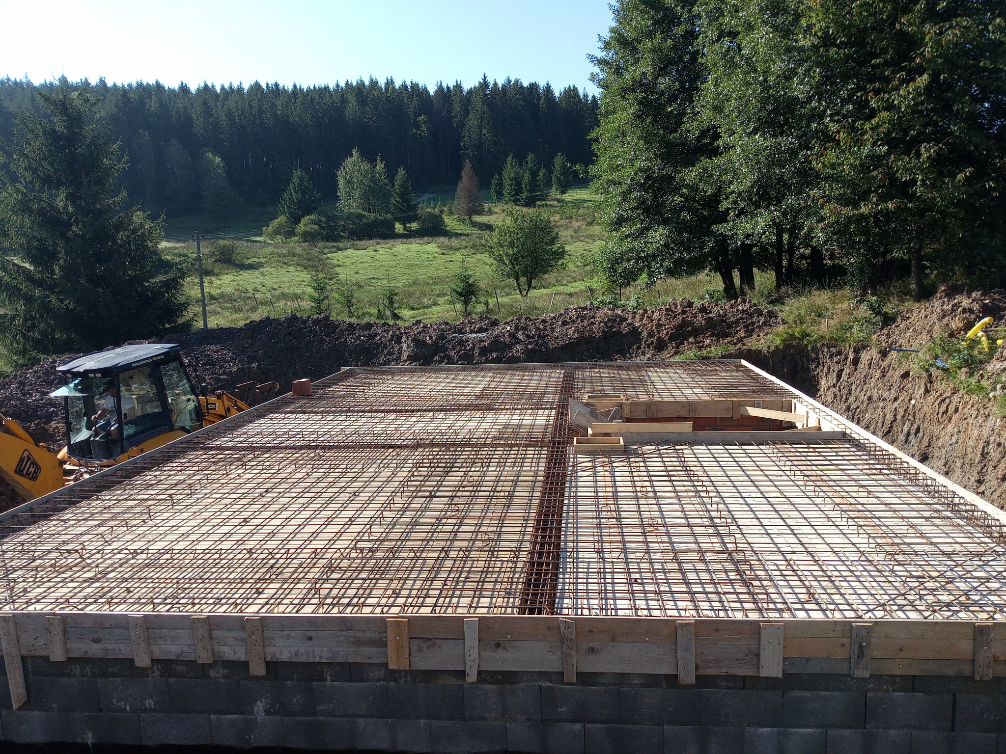 Zbrojenie fundamentu pod budowę domu jednorodzinnego, widok z góry. W tle las i łąka, po lewej koparka JCB. Konstrukcja z metalowych prętów i drewnianych szalunków.