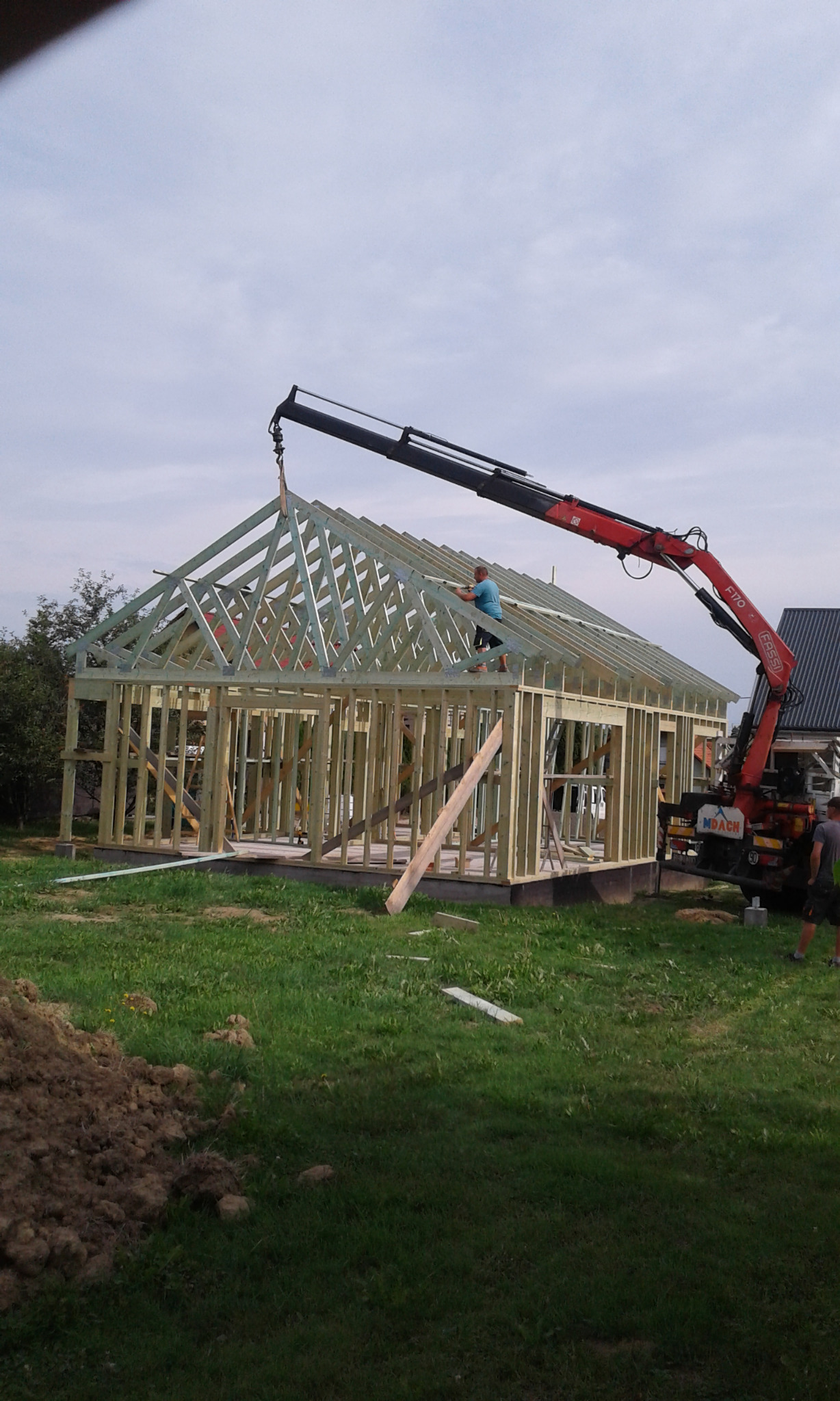 Budowa domu szkieletowego: montaż więźby dachowej z użyciem dźwigu. Pracownik na dachu, widoczne elementy konstrukcyjne i krajobraz działki.