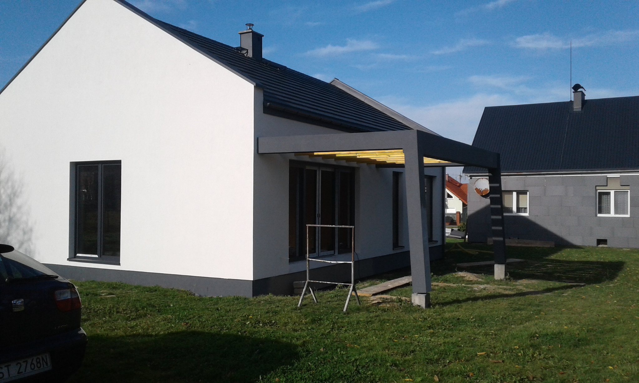 Nowoczesny dom z białą elewacją i szarą pergolą z żółtymi elementami, widok z zewnątrz na tle zielonego trawnika i błękitnego nieba. W tle drugi dom.