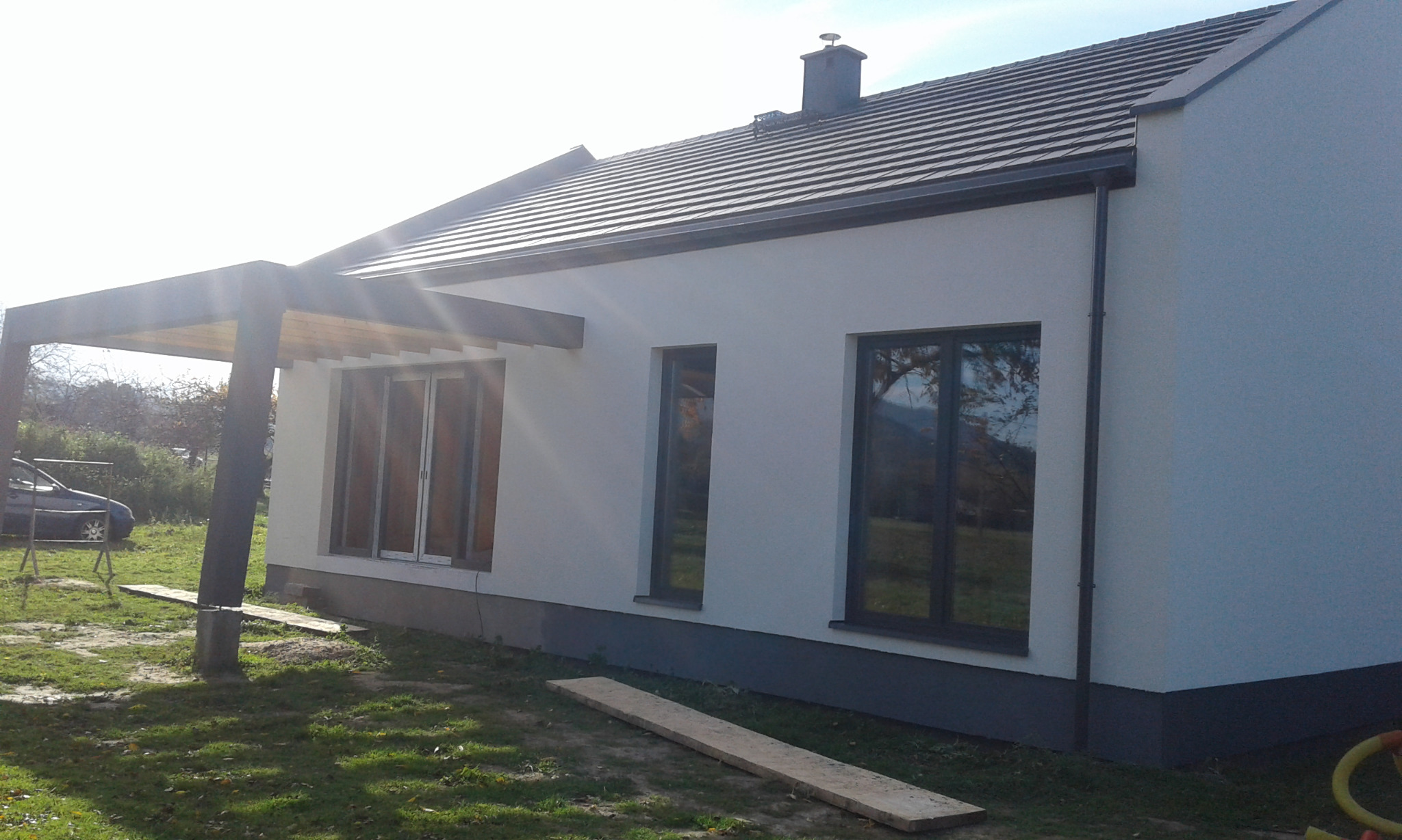 Nowoczesny dom z jasną elewacją, ciemną stolarką okienną i drewnianą pergolą. Widoczne detale wykończenia i otoczenie domu.