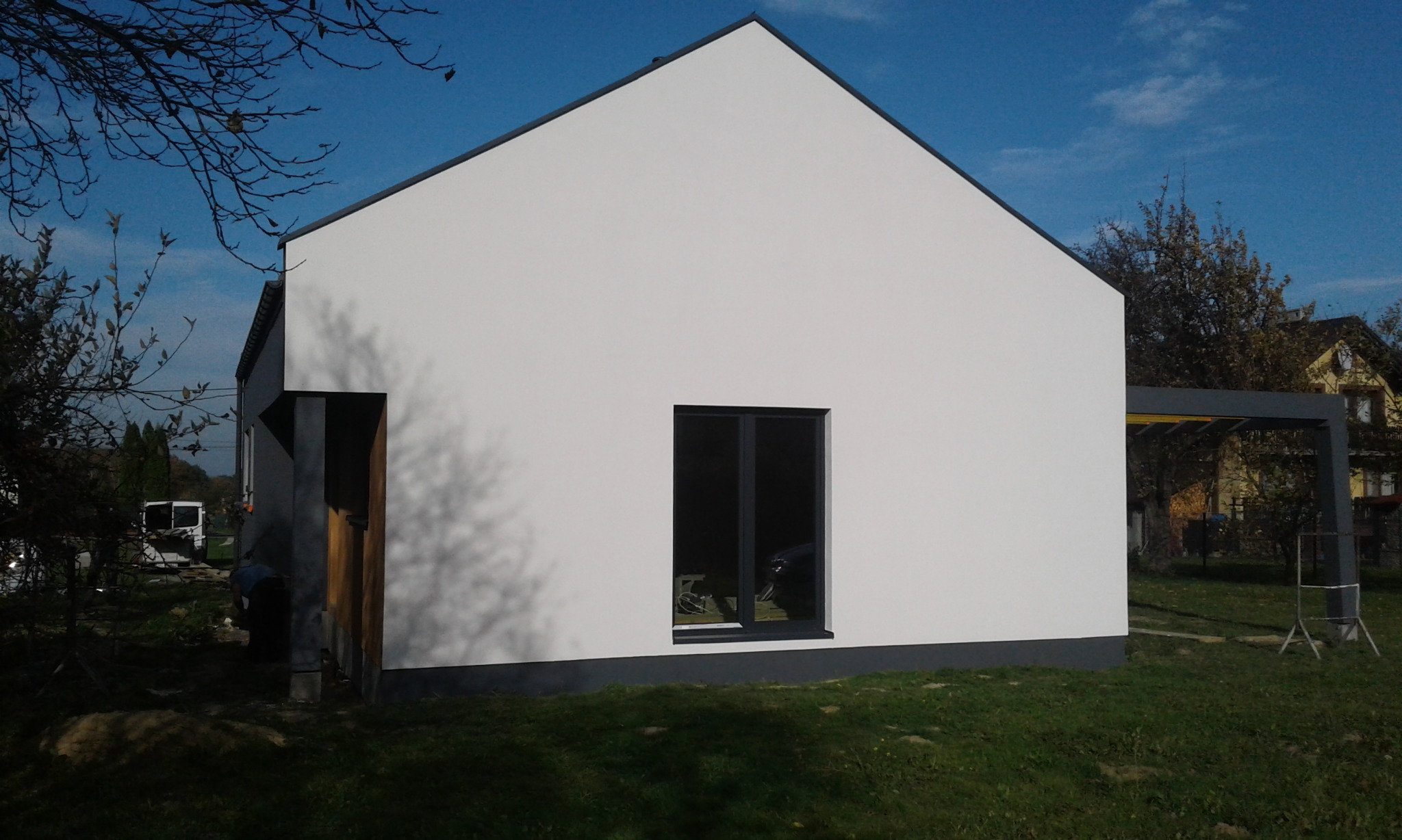 Nowoczesny, biały dom z szarymi akcentami, widok z zewnątrz. Minimalistyczna bryła, okno z ciemną ramą, fragment ogrodu i zadaszenia w tle. Świeżo otynkowana elewacja.