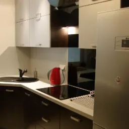 Nowoczesna kuchnia z ciemnymi szafkami dolnymi i jasnymi górnymi, czarnym zlewem i płytą indukcyjną, uzupełniona czerwonym czajnikiem i srebrną lodówką.