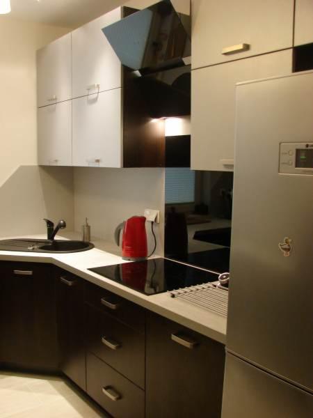 Nowoczesna kuchnia z ciemnymi szafkami dolnymi i jasnymi górnymi, czarnym zlewem i płytą indukcyjną, uzupełniona czerwonym czajnikiem i srebrną lodówką.