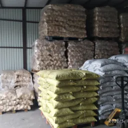 Magazyn z paletami worków brykietu drzewnego, niektóre owinięte folią, inne w kolorowych workach, widoczny wózek paletowy.